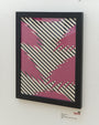 Framed artwork by Ryan De La Hoz showcases an abstract design of pink and black zigzags on diagonal black and white stripes, made with hand-cut paper, displayed on a white wall with a label in the bottom corner.