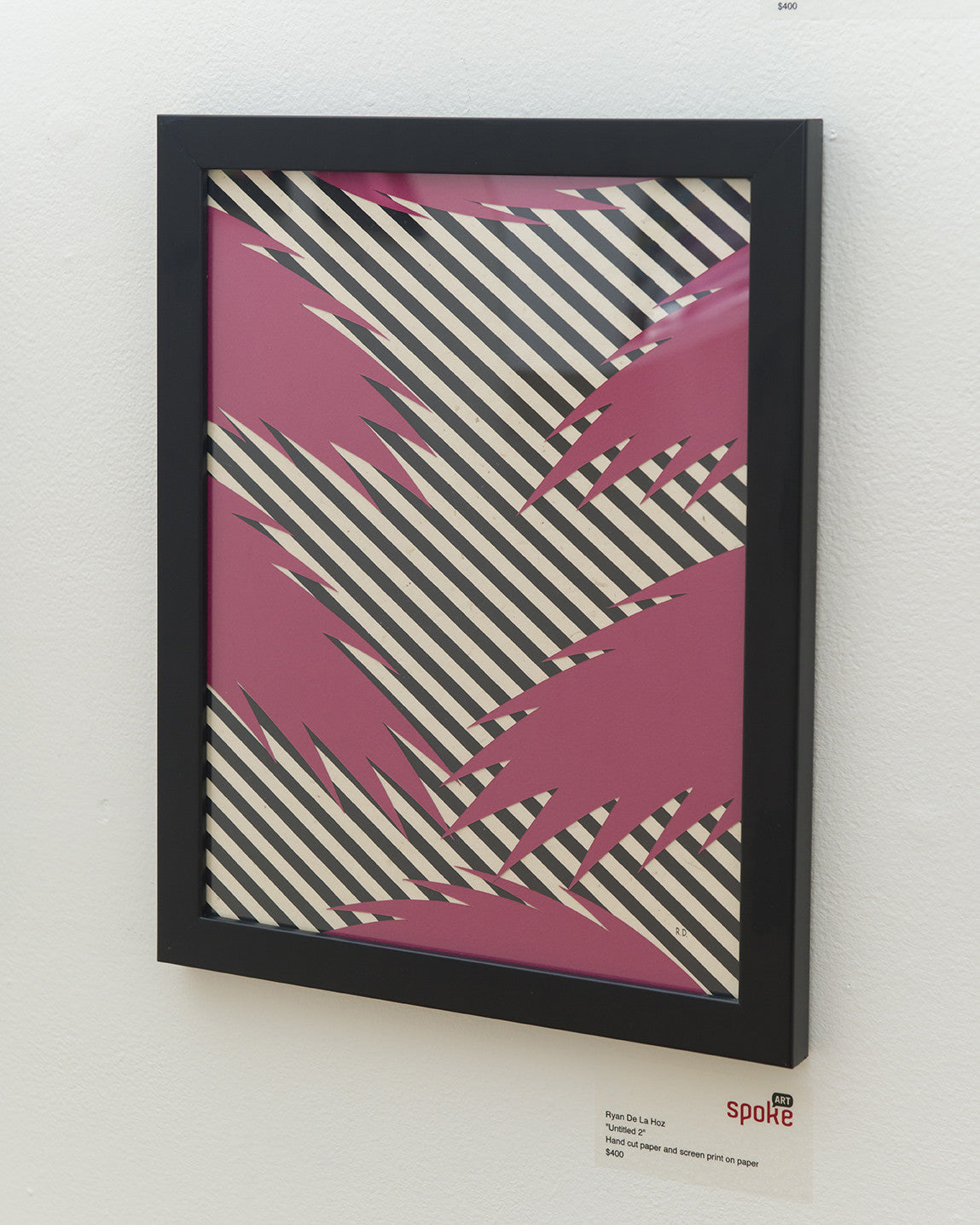Framed artwork by Ryan De La Hoz showcases an abstract design of pink and black zigzags on diagonal black and white stripes, made with hand-cut paper, displayed on a white wall with a label in the bottom corner.
