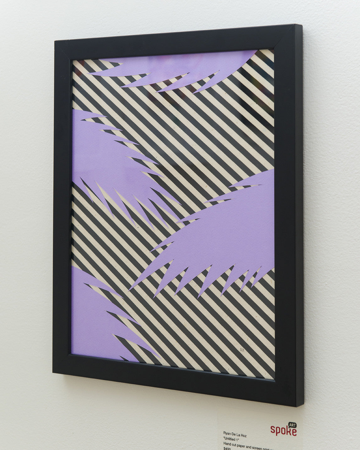 Framed black and white diagonal stripes are intersected by jagged purple shapes, crafted from hand-cut paper in a Ryan De La Hoz style. This exquisite piece is displayed in a black frame on a white wall with a small label beside it.