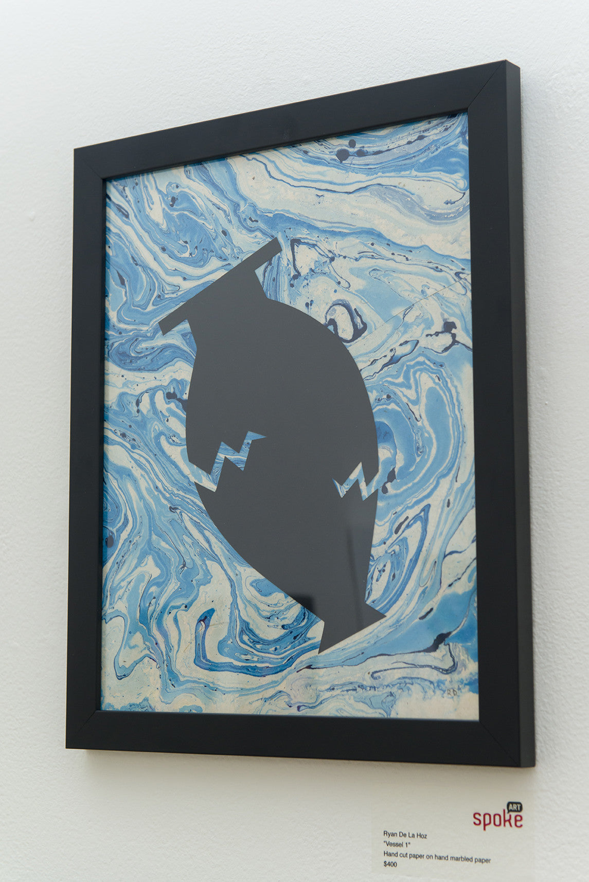 A Ryan De La Hoz piece, featuring a stylized grey vase with zig-zag lines on a marbled blue and white background crafted from hand-marbled paper, is displayed framed on a white wall with a small label.