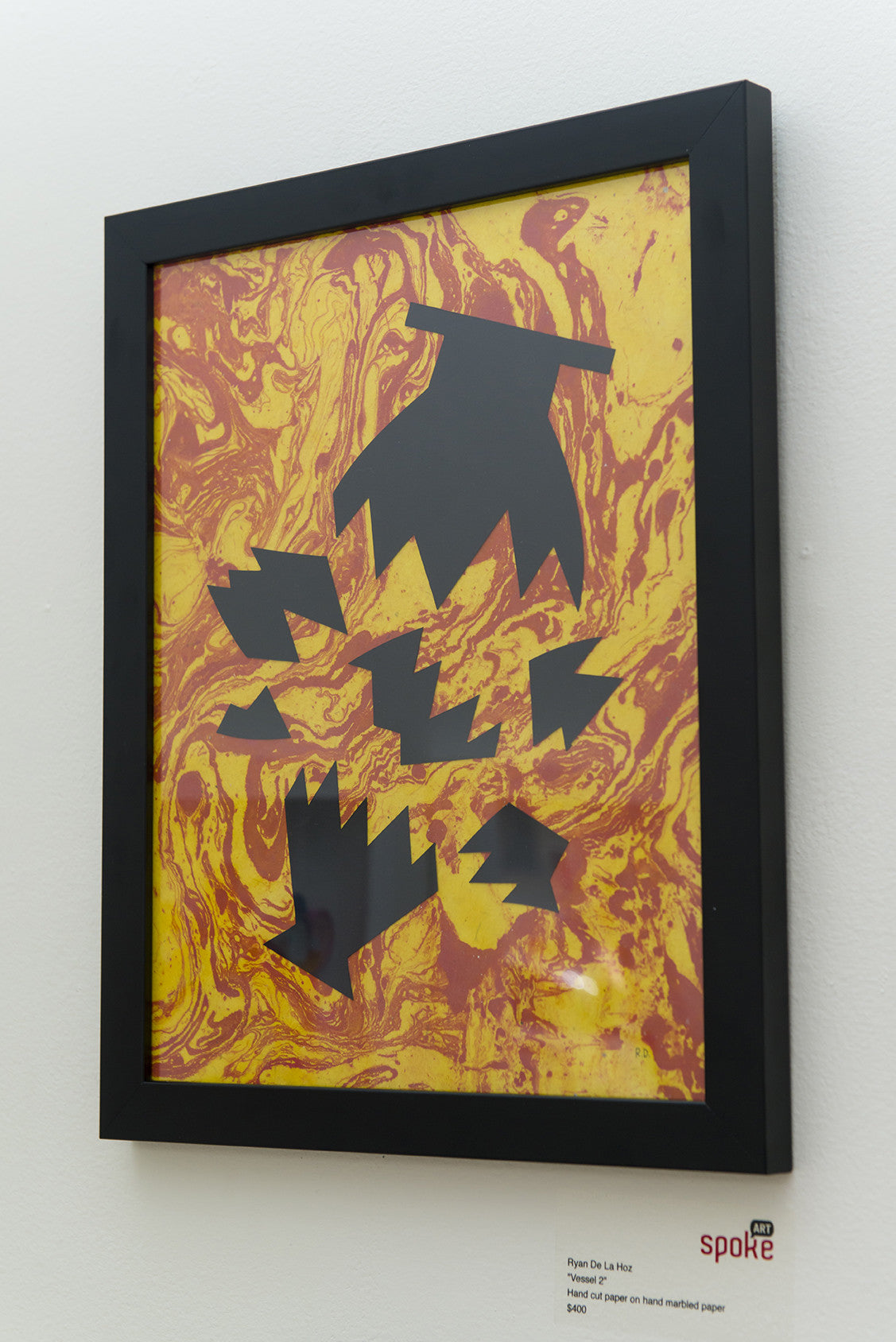 Framed abstract artwork titled Flynn De La Hoz, featuring black jagged shapes on a vibrant yellow and red hand-marbled background. Crafted with hand-cut paper, it echoes Ryan De La Hozs distinct style and is displayed on a white wall. Brand: Ryan De La Hoz.