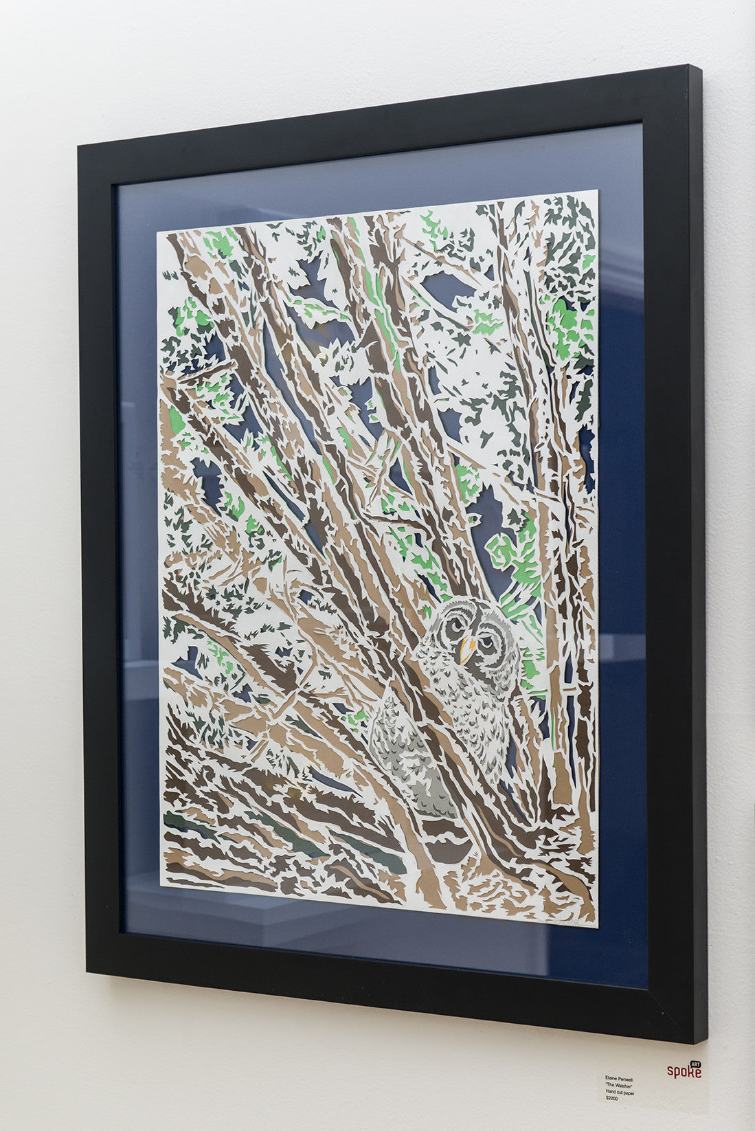 The Watcher by Elaine Penwell is a framed artwork featuring an owl perched in a tree with intricate branches and leaves. The hand-cut paper piece blends into the natural setting against a dark background, highlighting the owl’s delicate features and surrounding foliage.