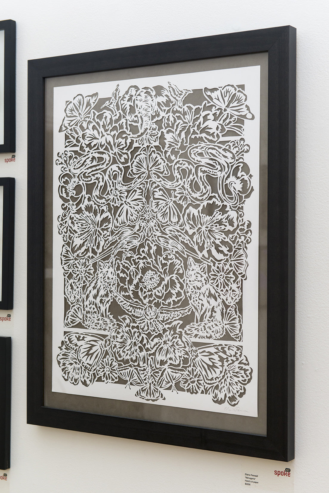 Elaine Penwells framed hand-cut paper art from the Menagerie Collection showcases exquisite floral and animal designs in predominantly white with black outlines, elegantly displayed on a wall.