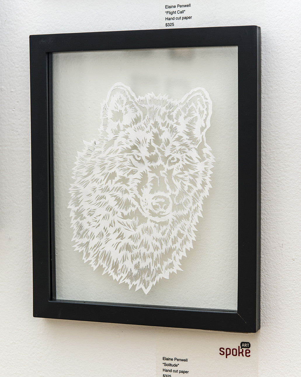Elaine Penwells framed artwork showcases a wolfs head intricately hand-cut from paper, symbolizing solitude. Mounted against a neutral wall, its black frame and gallery label highlight the artist and her work.