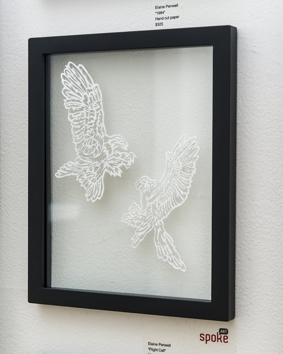 A framed art piece by Elaine Penwell showcases two intricately hand-cut paper birds titled Flight Call, facing each other with wings spread wide, set against a plain background within a sleek black frame.