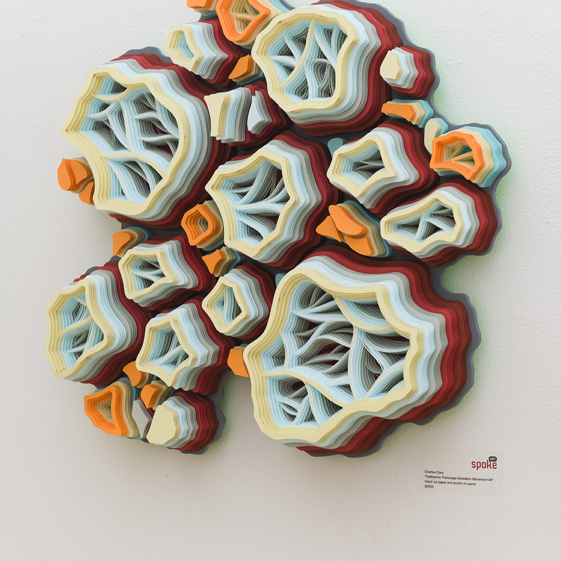 This vibrant 3D artwork by Charles Clary features layered, wavy shapes resembling abstract coral or geological forms. Hand-cut paper in orange, white, and blue adorns a white wall, with the artists signature in the corner.