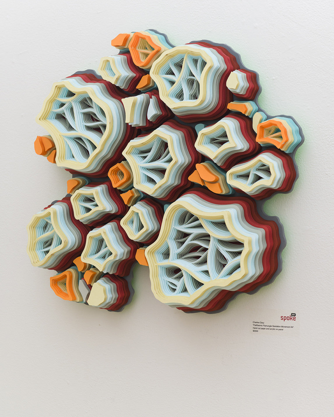 This vibrant 3D artwork by Charles Clary features layered, wavy shapes resembling abstract coral or geological forms. Hand-cut paper in orange, white, and blue adorns a white wall, with the artists signature in the corner.