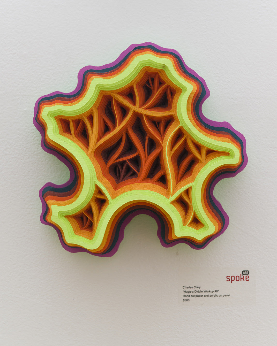 A layered sculpture by Charles Clary, resembling a stylized topographic map, showcases vibrant contours in yellow, orange, and purple. Crafted with hand-cut paper on acrylic panel, it casts intricate shadows against a pale wall. Text beside it includes the artists name and artwork details.