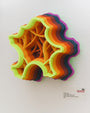 A 3D artwork by Charles Clary features layered, wavy concentric shapes crafted from hand-cut paper in neon green, orange, yellow, and purple. The piece resembles an abstract star or flower and is mounted on a white wall.