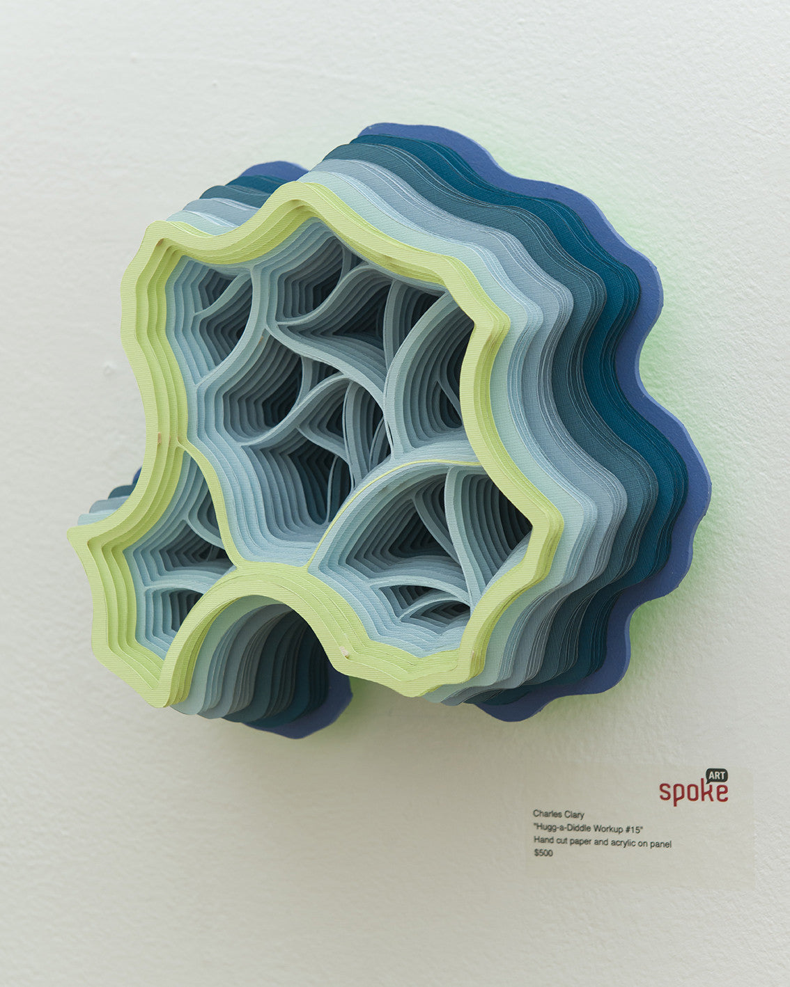 A Charles Clary hand-cut layered paper sculpture with wavy edges and shades of blue and green is mounted on a white wall, casting subtle shadows. It features an abstract, organic shape, made from acrylic on panel with descriptive text below.
