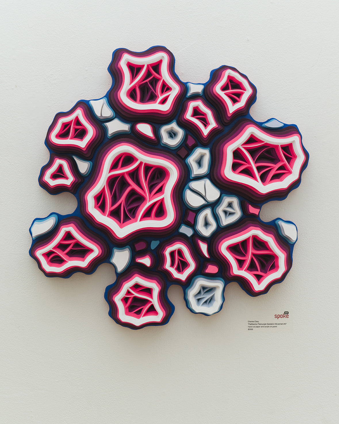 A Charles Clary-inspired abstract wall sculpture in pink, red, white, and blue features layered organic shapes resembling biological cells. The hand-cut paper art forms a 3D effect with interconnected cellular structures on a white background.