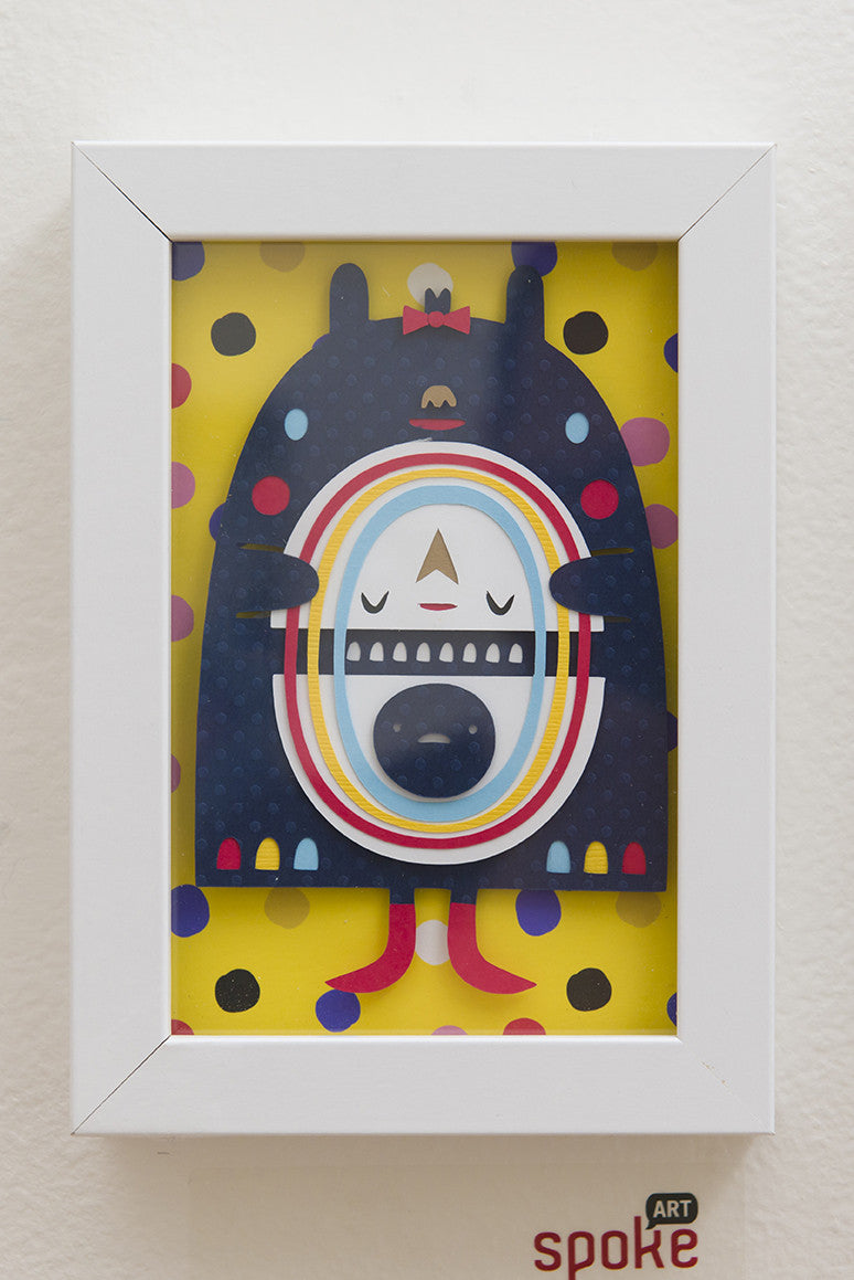 Crowded Teeth offers framed artwork featuring a Rainbow Monster with a round body, rainbow rings, closed eyes, and small limbs. Set against a yellow background with colorful dots, the piece is presented as a giclee print in a sleek white frame.
