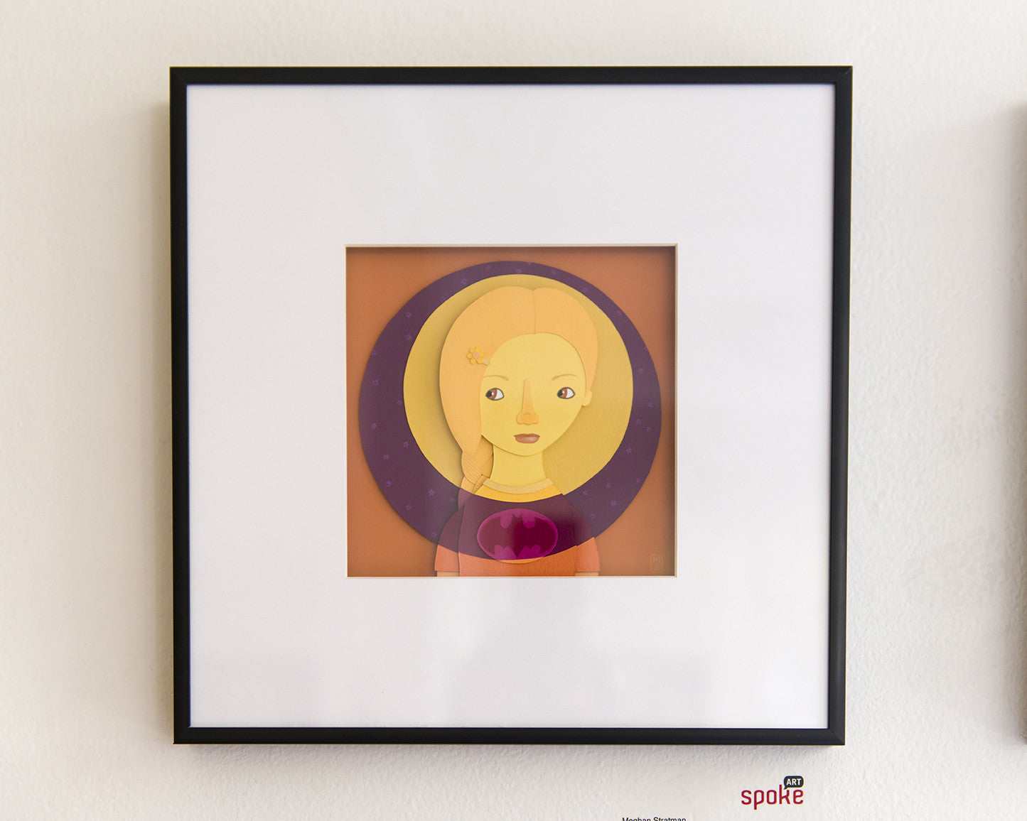 A framed artwork by Meghan Stratman depicts a stylized figure with yellow hair on an orange and purple circular background. Its minimalist style with bold colors and simple shapes is set in a black frame against a white wall, reflecting Stratmans signature paper collage aesthetics.