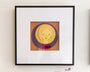 A framed artwork by Meghan Stratman depicts a stylized figure with yellow hair on an orange and purple circular background. Its minimalist style with bold colors and simple shapes is set in a black frame against a white wall, reflecting Stratmans signature paper collage aesthetics.