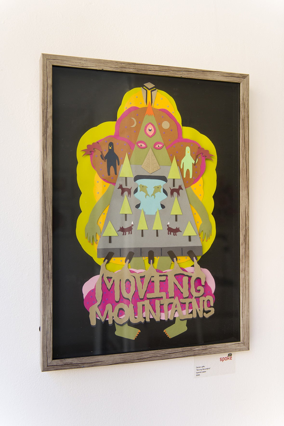 Framed Daniel Jaffe artwork features a colorful triangular mountain creature with eyes, trees, animals, and is crafted from layered paper for depth. MOVING MOUNTAINS adorns the bottom, enhancing its whimsical charm.