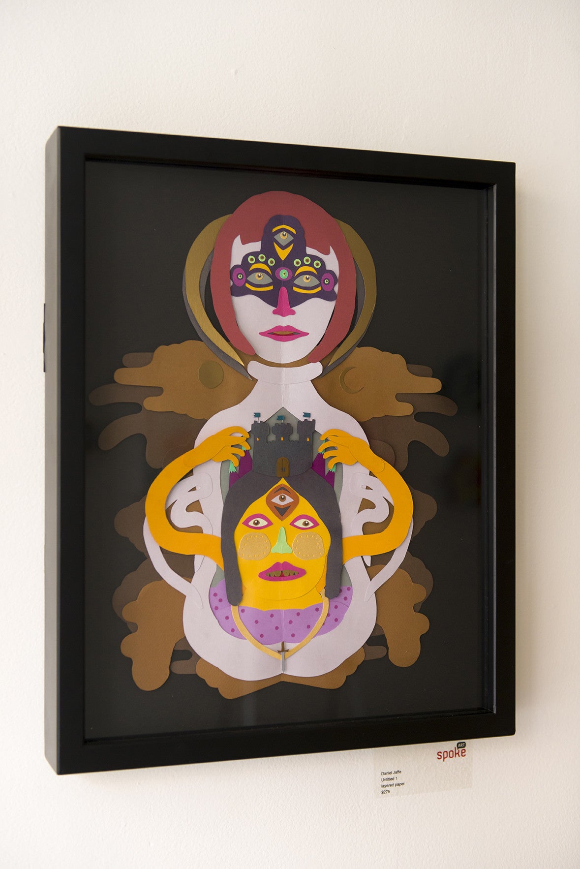 Framed abstract artwork by Daniel Jaffe showcases two human-like figures with colorful, symmetrical designs; the top features a multicolored face with closed eyes, while the bottom figure holds a head against a dark background with abstract shapes and layered paper effects.