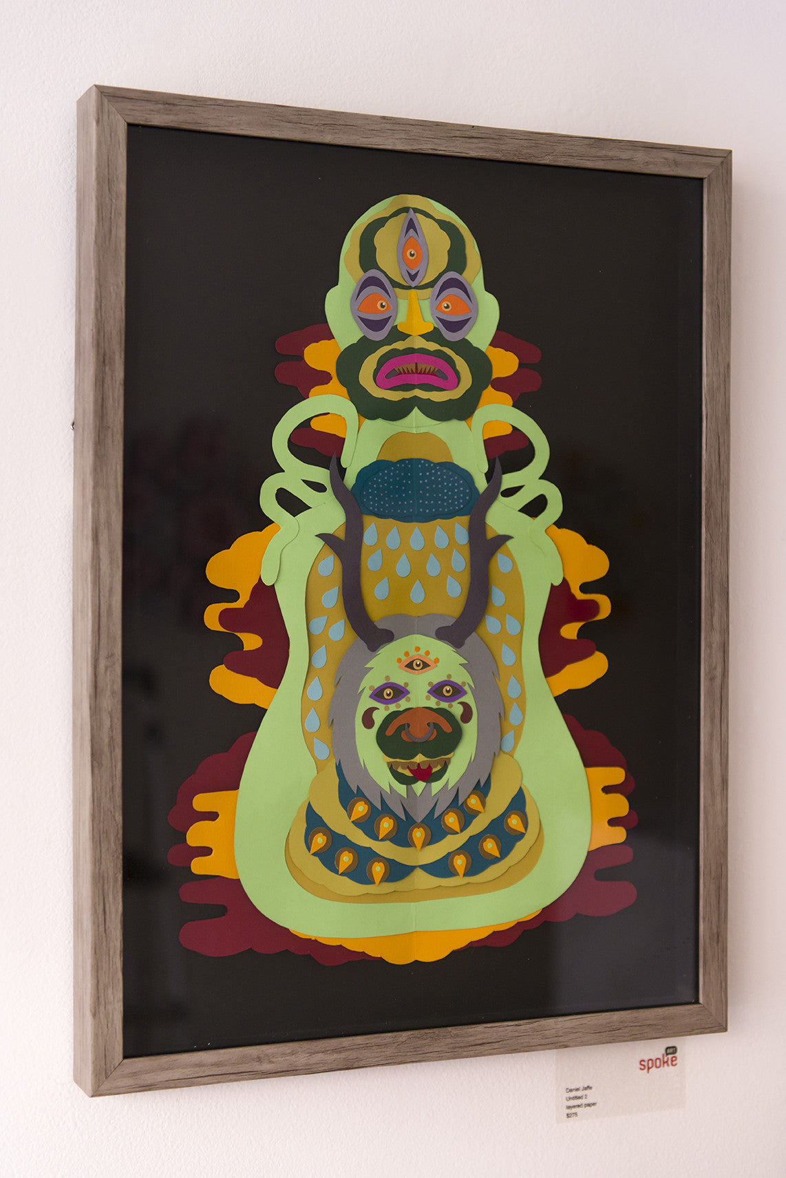 Untitled 2 by Daniel Jaffe is a framed abstract piece featuring vibrant, layered paper art with face-like shapes and eyes, adorned in green, yellow, and red patterns against a black backdrop on a white wall.