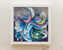 Crystal Wagners abstract art piece features swirling vibrant blues, purples, and greens that flow like waves or feathers. Framed in white, it showcases layered textures on a light background resembling an intricate screen print.