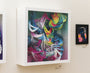 A lively 3D art piece with swirling, colorful abstract shapes, reminiscent of Crystal Wagners style, is set in a shadow box frame. It is surrounded by smaller framed works in relief print and screen print styles on a light-colored wall.