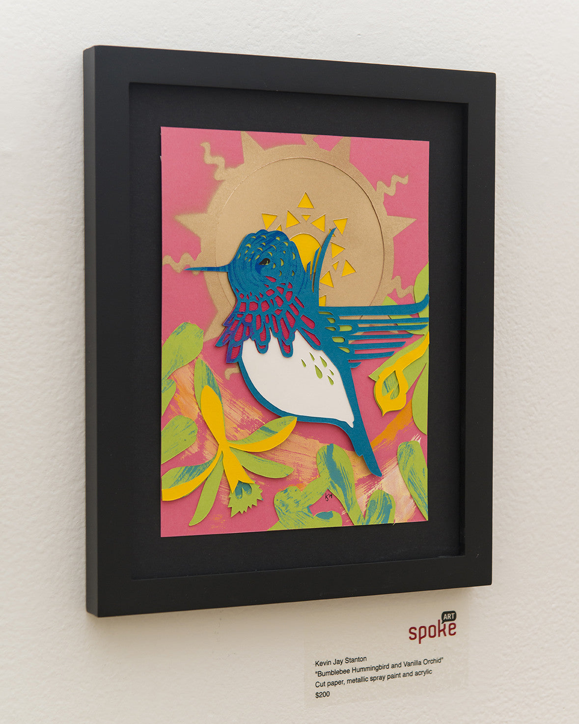 Kevin J Stantons framed artwork showcases a colorful abstract Bumblebee Hummingbird with blue and white feathers, surrounded by yellow flowers, a sun motif, and hints of Vanilla Orchid. The intricate cut paper art is displayed on a white wall.