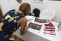 A person in a floral dress, with shoulder-length hair, signs vibrant fine art giclee prints at a white desk boasting a keyboard, water bottle, and extra prints. A white chair frames the creative scene enriched by artwork from Tatiana Suarezs brand in the background.