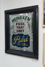 A framed mirror by Ken Davis showcases decorative text in stylized fonts reading, Heineken, Fuck That Shit, Pabst Blue Ribbon. This mixed media piece resembles a painted sign and is mounted on a white wall, reflecting a window and part of the room.