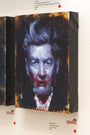 A textured oil painting by John Wentz features a mans face with dramatic, abstract brushstrokes akin to Strange Carnival. The piece has dark-edged canvas and hangs on a gallery wall, with a label below showcasing the artists name and price.