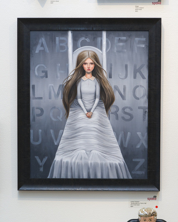 An Audrey Pongracz painting features a woman in a flowing white dress with long brown hair, standing before letter blocks. The muted gray backdrop complements this oil on linen board art titled, Tell me what you think of me.