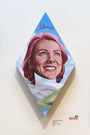 Johannah ODonnells diamond-shaped artwork in acrylic on wood portrays a woman with pink hair smiling against a sky and landscape, titled Silencio above her head. It includes a small label at the bottom right.