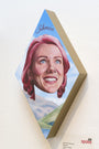 A diamond-shaped artwork by Johannah ODonnell shows a vividly painted woman with pink hair against a blue sky and landscape. It has Silencio at the top, crafted in acrylic on wood, and mounted on a light-colored wall.
