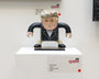 A male figure art sculpture with exaggerated features stands on a white pedestal at Spoke Art Gallery. Created by Geoff Trapp with Vinyl Sculpy, it wears a suit and tie, featuring black arms and cream-colored hands, marked as part of an exhibit.