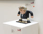 A cartoonish vinyl Sculpy figure from Geoff Trapp with exaggerated features stands on a display pedestal at the Spoke Art Gallery, wearing a suit and gray hair. A partially visible exhibition label is accompanied by a red sticker on the wall.