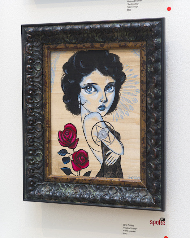 A stylized woman with big eyes and short black hair, tattooed with a steering wheel, anchor, and two red roses as an homage to Dorothy Vallens, is showcased in an acrylic painting by Sandi Calistro at Spoke Art Gallery. It features a dark ornate frame.
