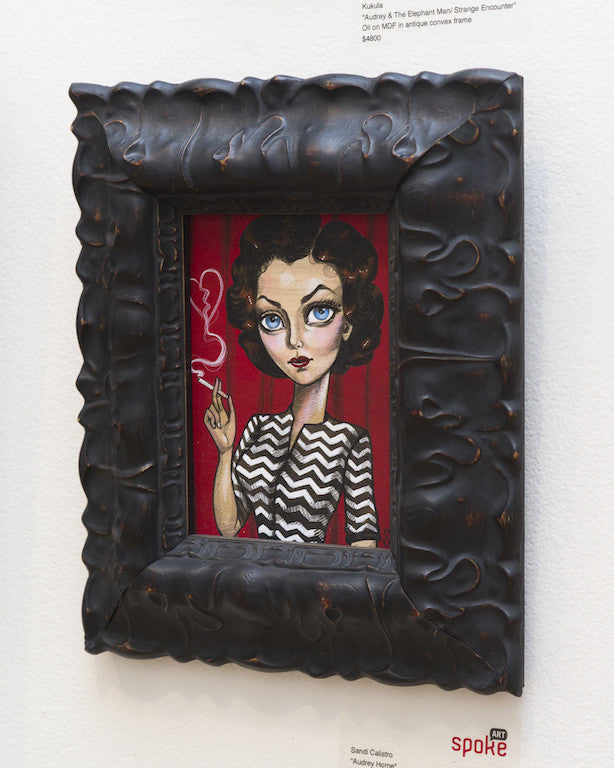 A painting in a carved wooden frame from Sandi Calistro portrays a woman with stylized features and large eyes, holding a cigarette. She wears an Audrey Horne-esque black-and-white zigzag dress against a red background. The dark frame is ornate.