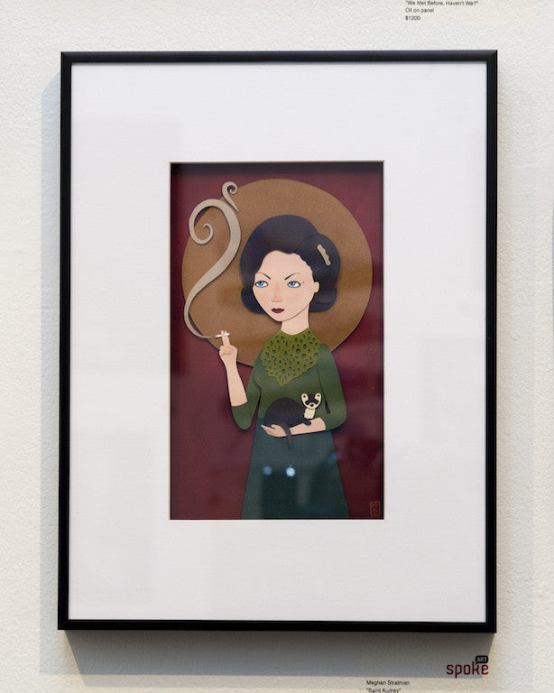 Framed artwork by Meghan Stratman features a stylized dark-haired woman in a green dress holding a cigarette with swirling smoke and a small dog, set against a red and gold background that enhances the paper collage elements, all in an elegant gallery setting.