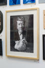 A black and white portrait by Joel Phillips depicts a serious man holding a chicken. Created in charcoal and graphite, this framed artwork is displayed on a gallery wall among captivating pieces.