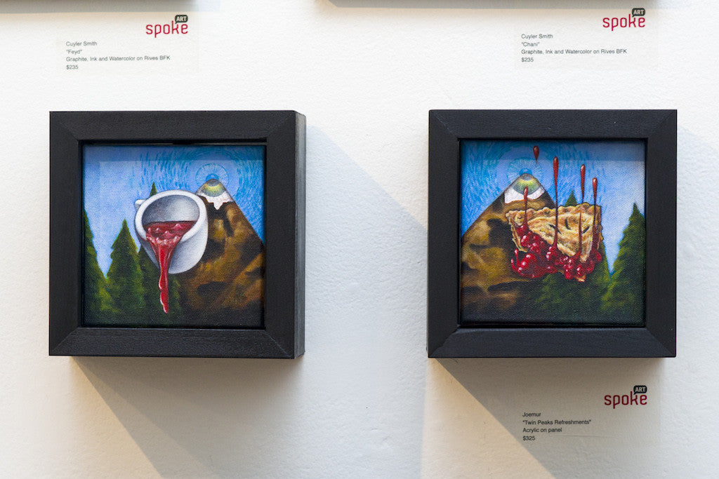 Two small framed paintings of mountain landscapes in acrylic hang on the wall. Joemurs piece on the left depicts a cup pouring red fluid, while Frank Emerges work features dripping red liquid like jam or blood. Each frame is crowned with an artist label.