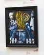 Frank Emerges, an acrylic on panel by Joemur, shows a surreal scene with a fiery-headed figure and flaming ear. The phrase Pab Blue floats above a stern dark blue face. The framed painting hangs mounted on a gallery wall.