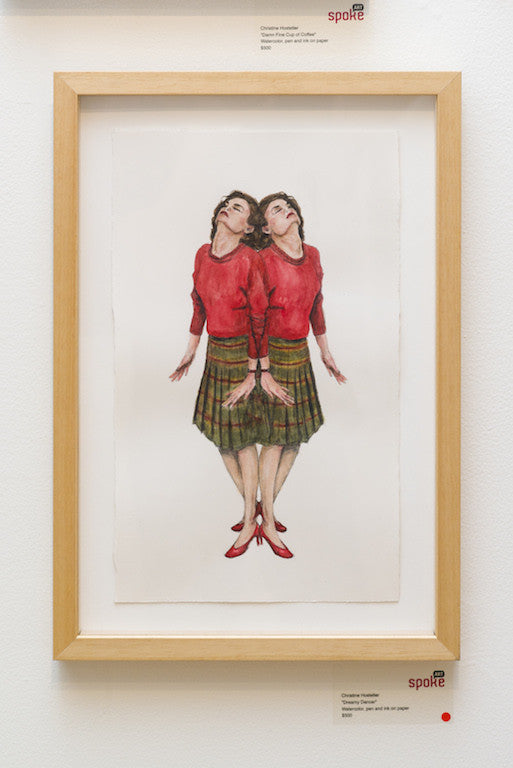 A framed watercolor artwork by Christine Hostetler shows two women in mirrored poses, back-to-back. They wear matching red tops, plaid skirts, and red shoes with heads tilted upward. The plain background enhances the dreamy dancers elegance in this piece from Spoke Art Gallery.