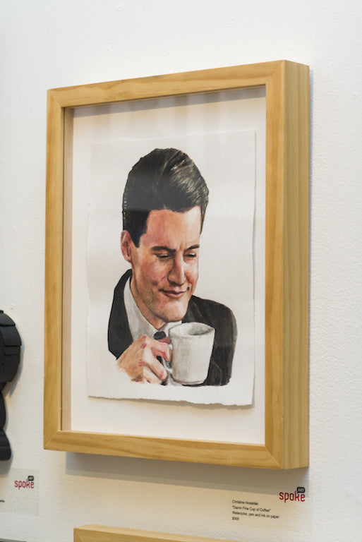 A watercolor painting by Christine Hostetler depicts a man in a suit, enjoying a fine cup of coffee with a content expression. It is elegantly framed in wood and subtly labeled beneath on a white wall.