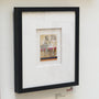 A black-framed artwork with a white mat on the wall showcases a seated figure amidst colorful elements. A label reveals its Graphite on found matchbook by Jason DAquino, enhancing its allure.