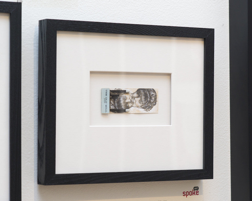 Framed artwork featuring a detailed illustration of a woman with stylized hair on currency-like paper, reminiscent of Jason DAquinos style. The piece resembles graphite on found matchbook covers, centered in a white mat and encased in a black frame against a white wall. Brand: Jason DAquino.