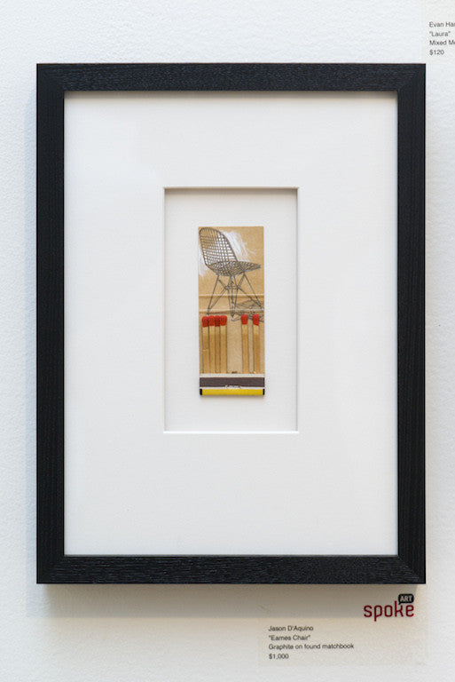 This artwork by Jason DAquino features a graphite drawing of an Eames-style wire chair on a matchbook, with red-tipped matches at its base. Its perfectly centered in white matting and enclosed in a sleek black frame.
