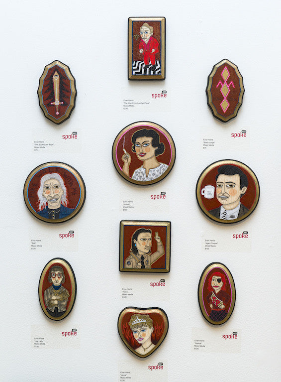 On a wall, nine framed paintings by Evan Harris showcase stylized portraits and geometric designs nodding to the Log Lady. Varying in shape and size, they feature diverse characters in red, black, and white tones. Each piece is labeled spoke.