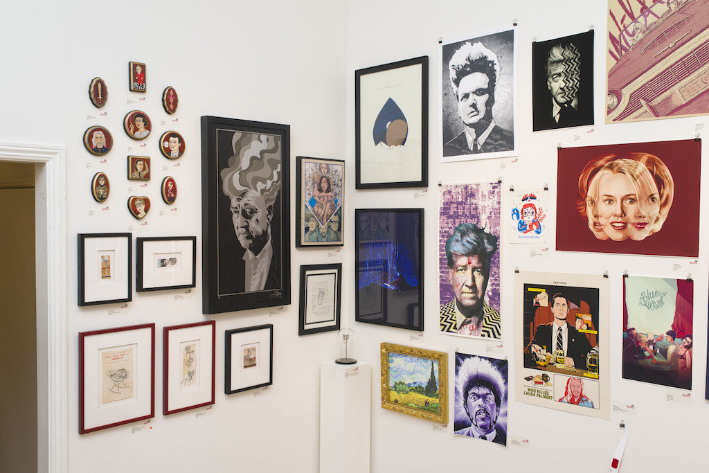 A gallery wall showcases an eclectic mix of fine art, featuring portraits, abstract designs, and landscapes. Frames and styles vary, creating a vibrant display on a white wall that includes a Sam Gilbey giclee edition for added flair.