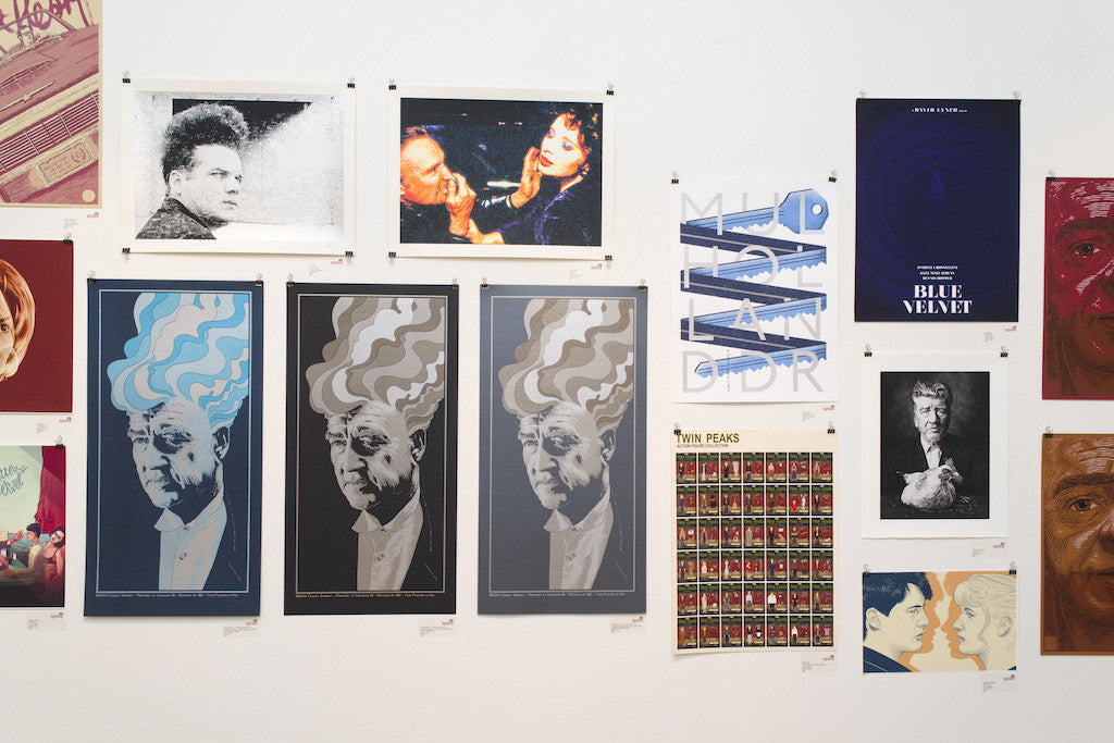 The wall display by Matt Weller showcases fine art giclee prints and posters, featuring abstract portraits with wavy patterns, Blue Velvet-themed artworks, and a grid of twin images. The color palette includes blues, reds, and earth tones.