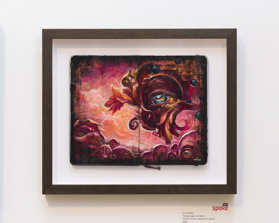 NC Winters framed artwork Dreamscape In E Minor is a surreal, vibrant acrylic painting with a central eye and swirling abstract shapes in red, pink, and orange. Displayed on an open notebook against a white wall.