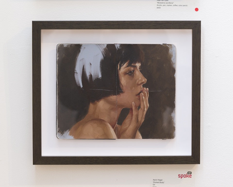 Aaron Nagels framed oil portrait on Moleskine captures a woman with short dark hair, thoughtfully holding her hand to her mouth. This exquisite piece is beautifully displayed on a white gallery wall.