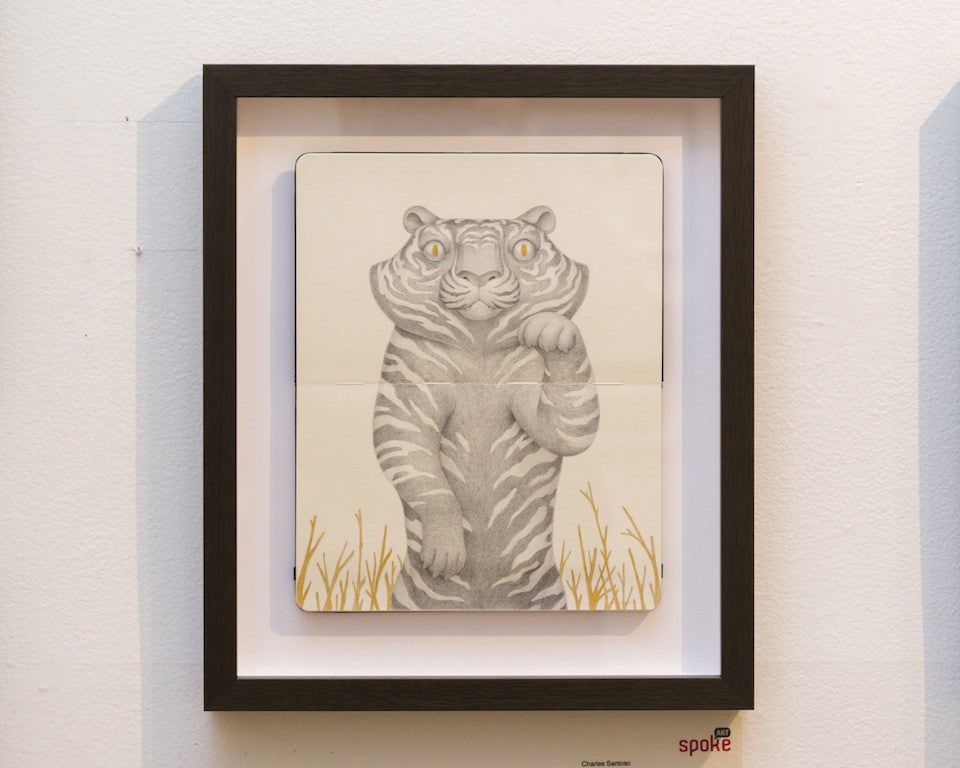 Charles Santosos framed artwork showcases a Black Lucky Tiger on its hind legs. Expertly drawn in pencil and gold ink, the tigers yellow eye contrasts against a white backdrop encircled by golden grasses. The art is elegantly framed in black.