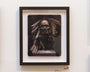 A framed artwork by Greg Gandy features an Ancestor Skull with feathers and beads against a dark background. The black frame with a subtle white border enhances this evocative piece, displayed on a white wall.